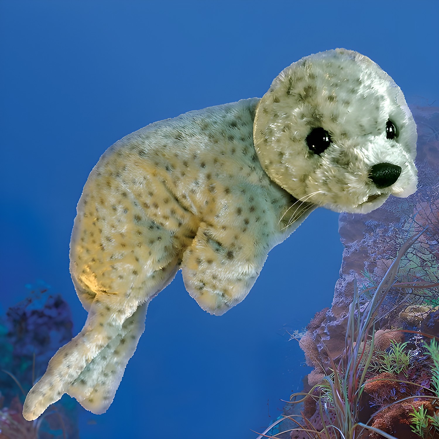 Folkmanis harbor seal baby hand puppet in light grey fur with dark spots, black glass eyes and whiskers, against a blue ocean background
