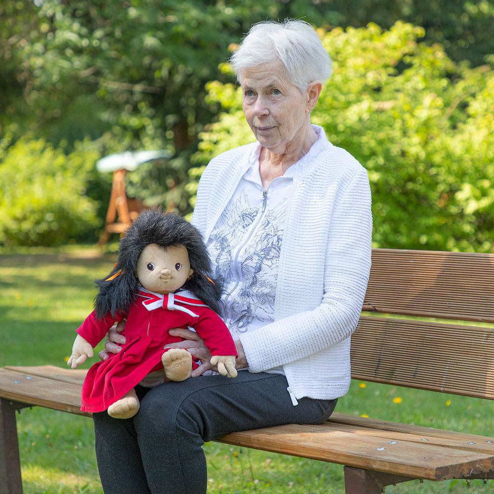 Seniorin mit der Joyk Empathiepuppe Mia auf dem Schoß auf einer Gartenbank