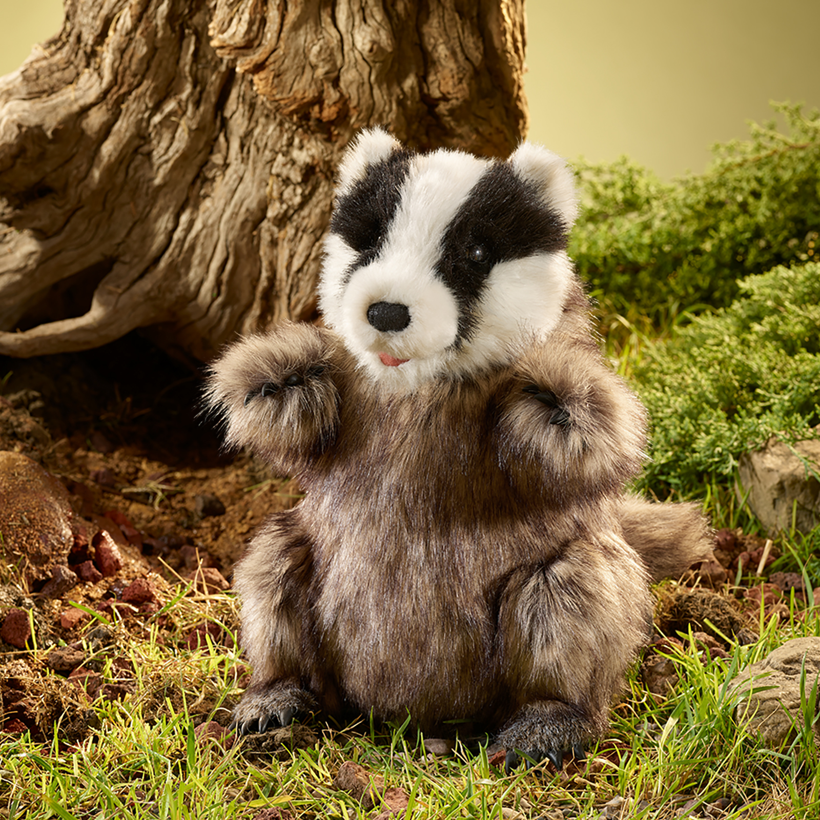 Folkmanis badger hand puppet sitting in grass by a tree trunk, with its striped head and mouth slightly open.