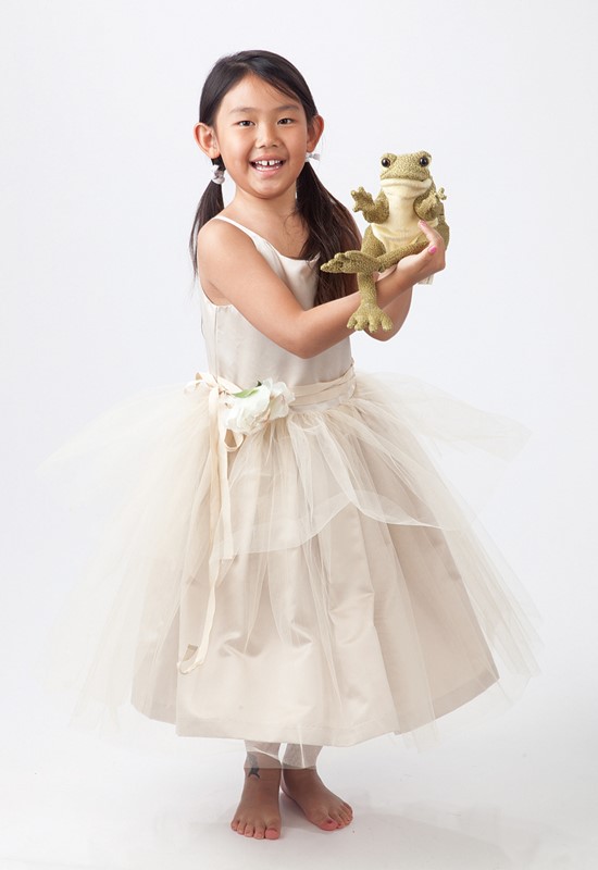 Girl in a cream tulle dress holding the Folkmanis Funny Frog hand puppet in olive-green plush up to the camera