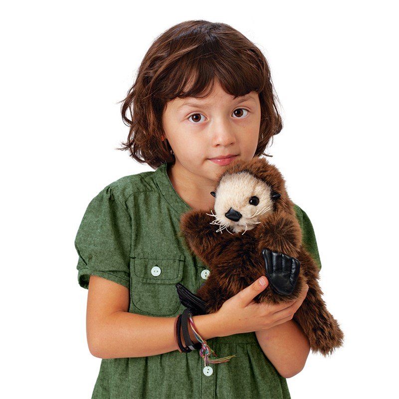 Girl holding a Folkmanis baby sea otter hand puppet with dark brown fur and black flippers against a white background
