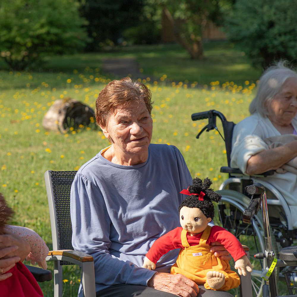 Seniorin mit der Joyk Empathiepuppe Naomi auf dem Schoß bei einer Gartenrunde