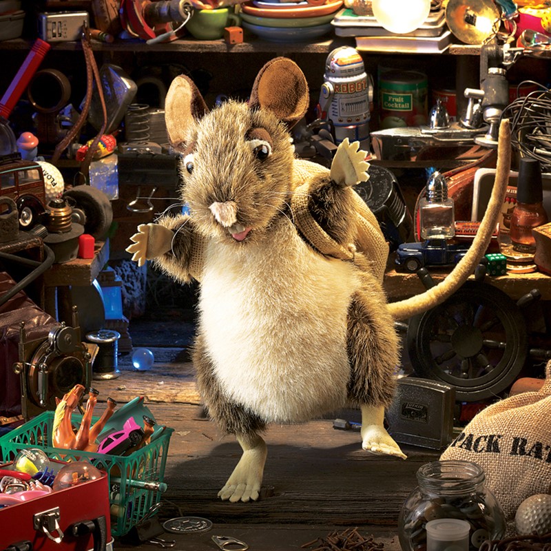 Folkmanis Pack Rat hand puppet with grey-brown fur and burlap sack standing upright in a scene full of found objects