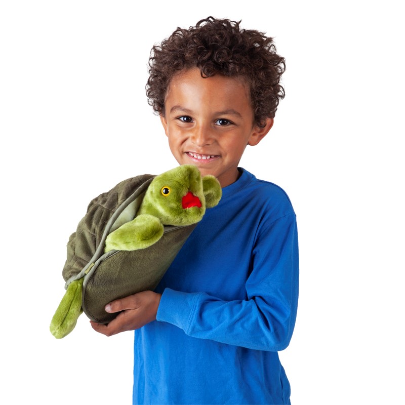Boy in blue holding the green Folkmanis Turtle hand puppet with red mouth against his arm, front view.