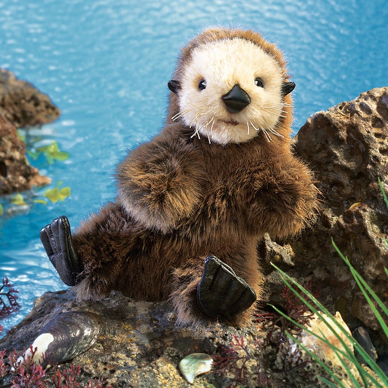 Folkmanis baby sea otter hand puppet with dark brown fur and a cream-coloured face, sitting on rocks in front of blue water