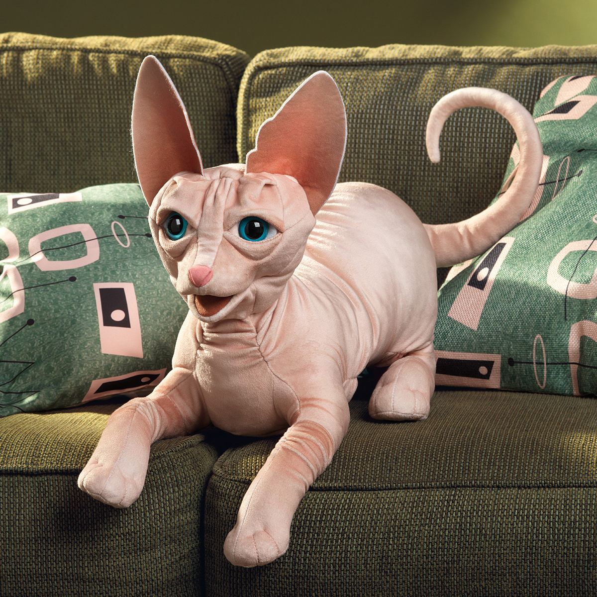 Folkmanis Handpuppe Sphynx Katze in Rosa-Beige mit türkisfarbenen Augen und großen Ohren, liegend auf grünem Sofa
