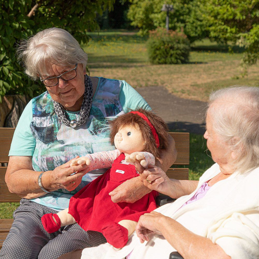 Two senior ladies looking at the Joyk empathy doll Ingrid together on a garden bench