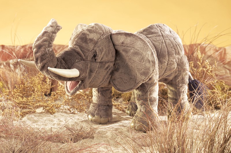Folkmanis elephant hand puppet in grey plush with raised trunk, open mouth and white tusk, standing in front of a savannah backdrop