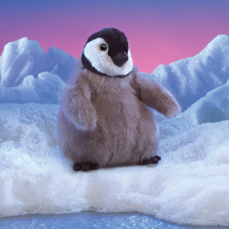 Folkmanis hand puppet baby emperor penguin with grey-brown down, black cap and white face, standing on snow in front of an ice backdrop