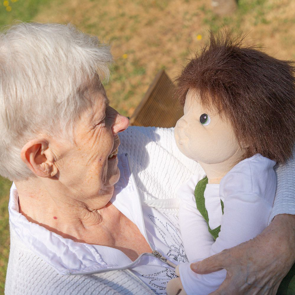 Senior lady making eye contact with the Joyk empathy doll Marco in the garden