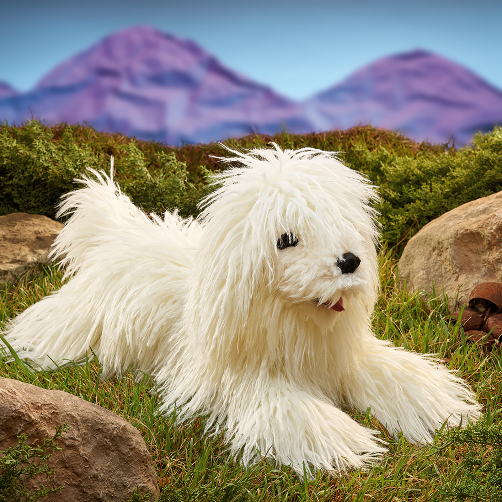 Folkmanis Komondor hand puppet lying among rocks in the grass, its mouth open.
