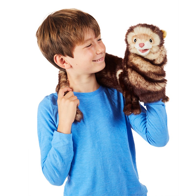 Boy holding a Folkmanis ferret hand puppet with brown fur and open mouth on his right hand, tail draped over his shoulder