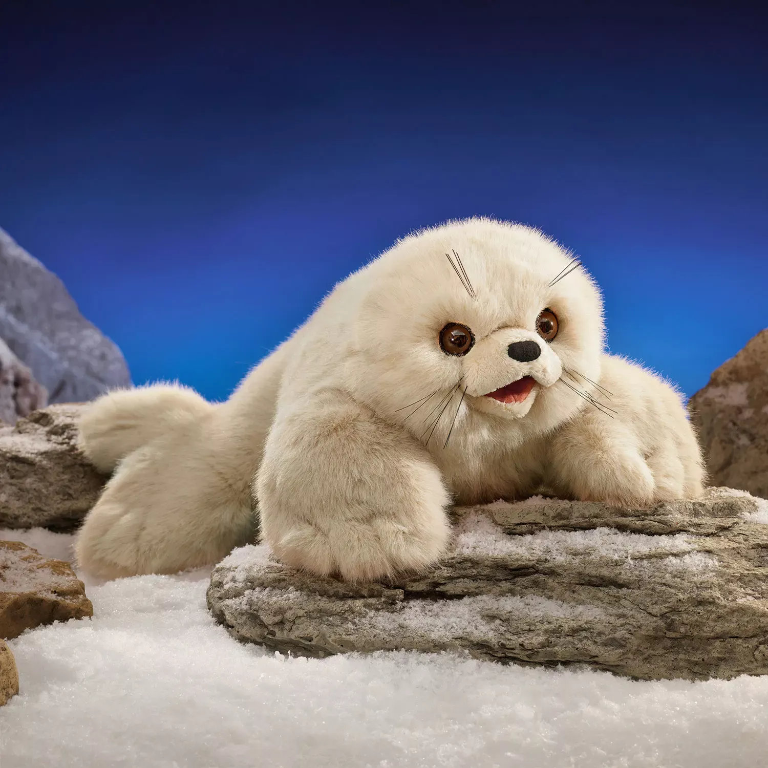 Folkmanis Handpuppe Sattelrobben-Baby mit cremefarbenem Plüschfell, geöffnetem Maul und Schnurrhaaren auf schneebedeckten Felsen
