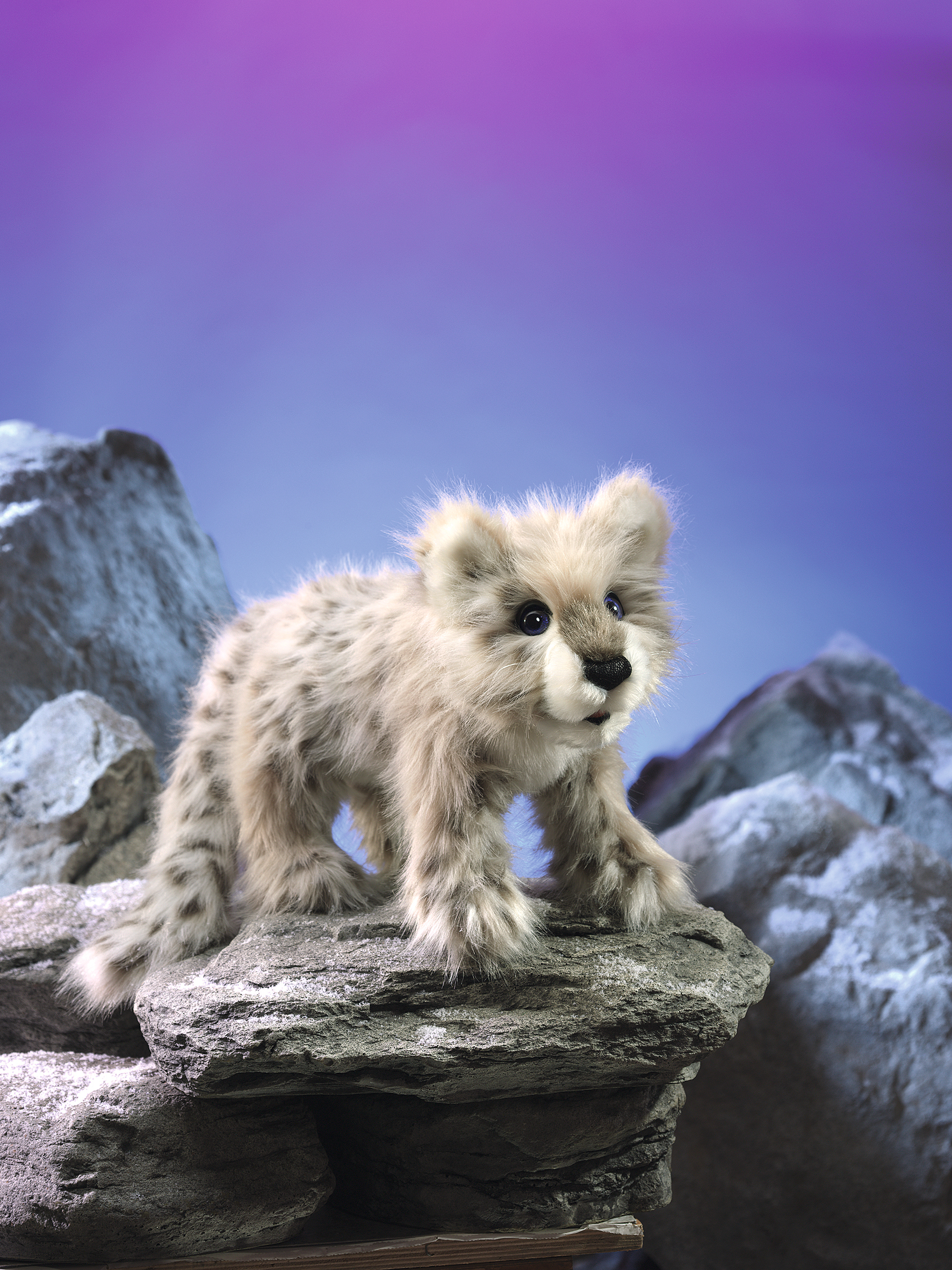 Folkmanis Handpuppe Baby Schneeleopard mit grau-beigem Fleckenfell, stehend auf einem Felsen vor blau-violettem Hintergrund